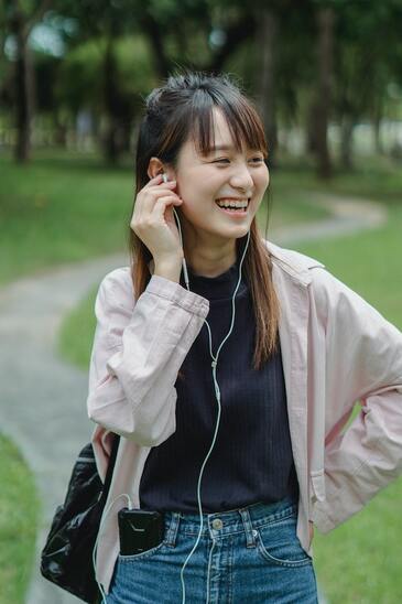 A woman is smiling as she walks along a path in a green park, talking on the phone using a set of earphones