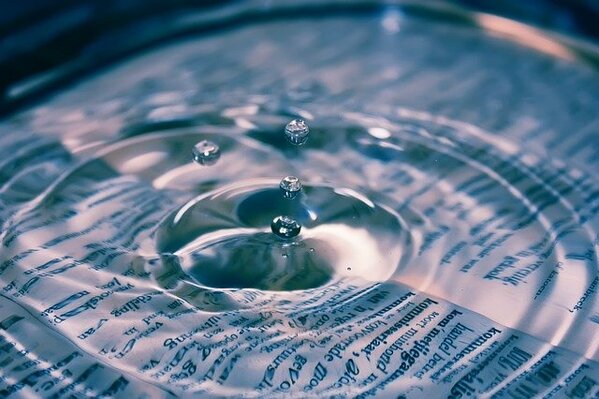 A transparent pool of water on top of a white document with printed text on it. A drop is falling into the pool of water and causing a ripple effect.