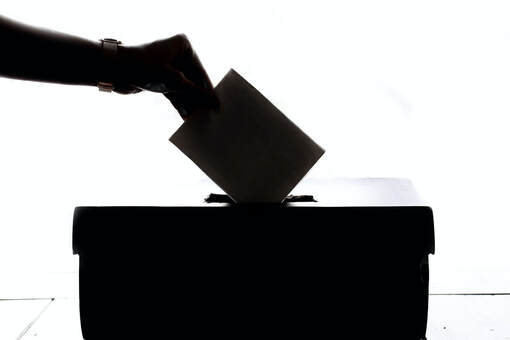 An election ballot box, with an arm reaching over it to drop a voting slip inside. Everything is in shadow so the photo is mainly dark colours.